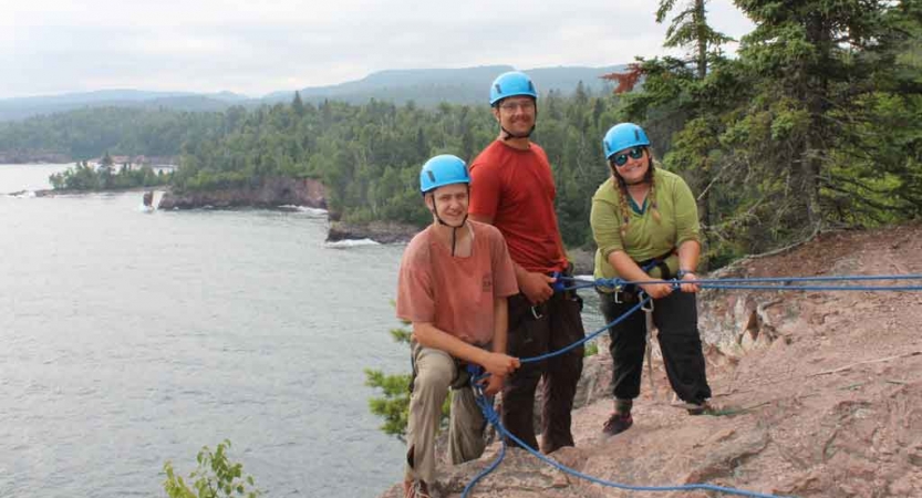 Three people wearing safety gear and secured by ropes stand on a rocky cliff and smile at the camera. They are high above a lake. 